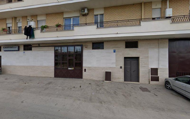 Vista stradale di un edificio beige a più piani con una porta d'ingresso marrone scuro, diversi balconi con piante in vaso e un'auto parcheggiata sulla destra. L'edificio ha un cartello con la scritta "CARPINELLI.