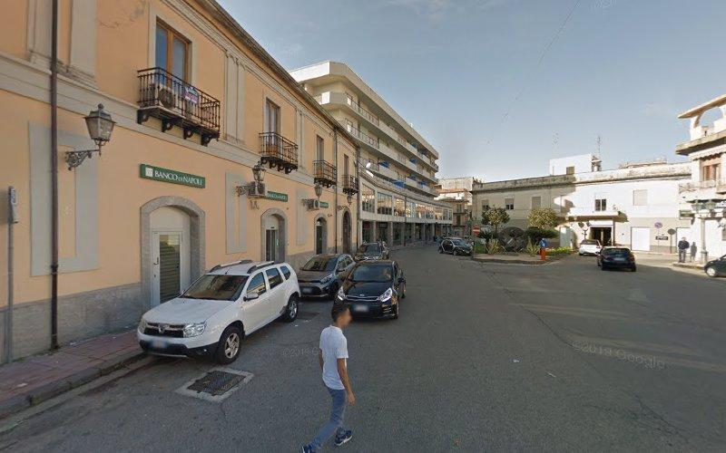 Una persona cammina su una strada con auto parcheggiate di fronte a un edificio a più piani con balconi. Un cartello di una banca (Banco di Napoli) è visibile sull'edificio a sinistra.