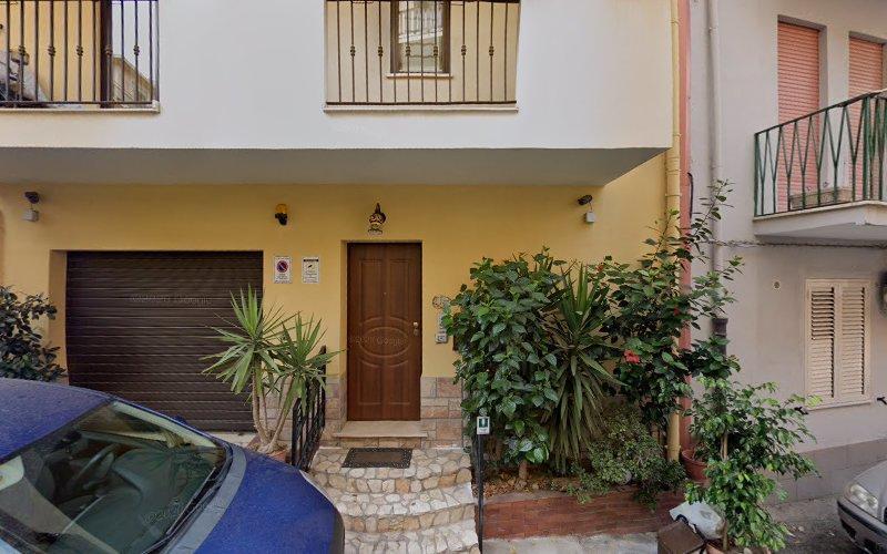 Una casa a due piani con facciata gialla, porta d'ingresso in legno e porta da garage nera. Piante verdi costeggiano il vialetto d'ingresso. Un'auto blu è parcheggiata di fronte. Gli edifici adiacenti sono visibili su entrambi i lati.