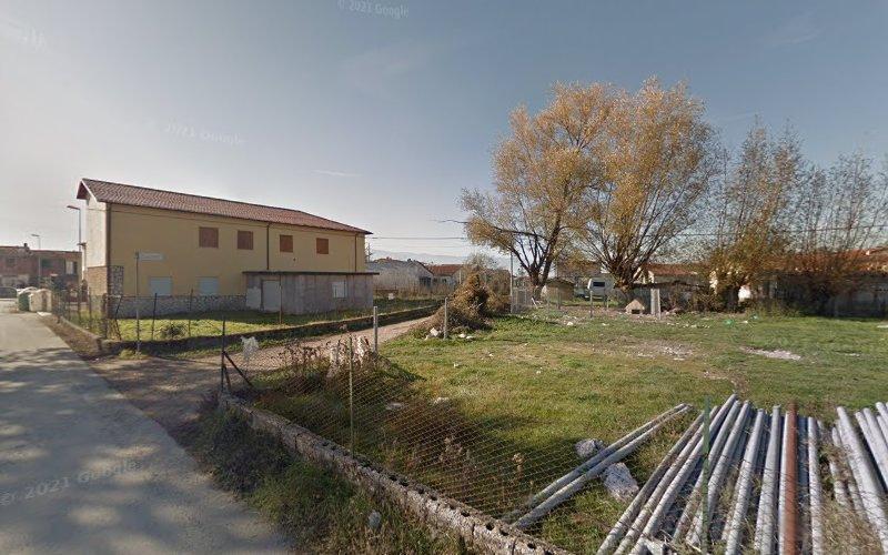 Un lotto vuoto con alcuni tubi sparsi e detriti, delimitato da una recinzione, accanto a un edificio residenziale e una strada. Alberi e altre case sono visibili sullo sfondo.