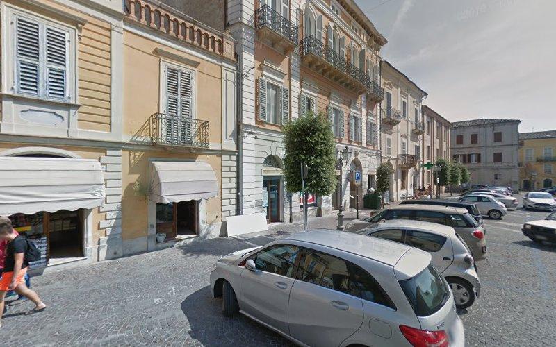 Una scena di strada che mostra auto parcheggiate di fronte a edifici a più piani con vetrine e balconi. Le persone camminano sul marciapiede e gli alberi sono piantati lungo la strada.