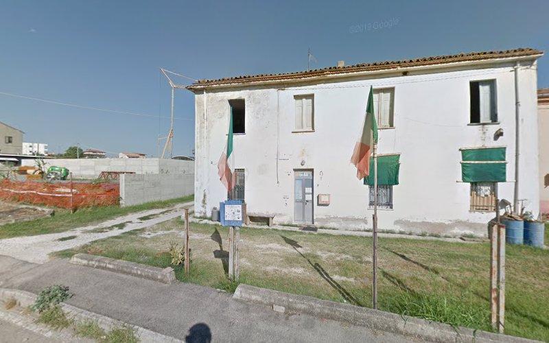 Un edificio bianco a due piani con due bandiere italiane esposte all'esterno. La struttura sembra invecchiata, con qualche segno di usura visibile. L'area circostante ha un marciapiede di ghiaia e materiali da costruzione nelle vicinanze.