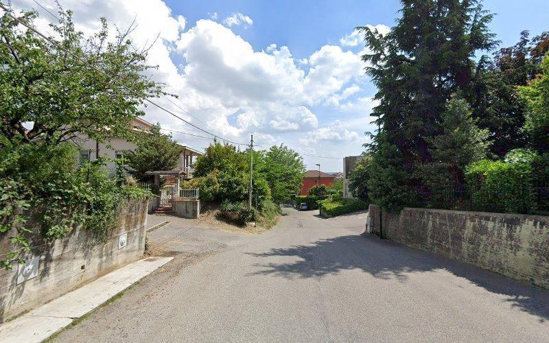 Una tranquilla strada residenziale con verde su entrambi i lati, una casa con un portico sulla sinistra e un muro di cemento sulla destra, sotto un cielo parzialmente nuvoloso.