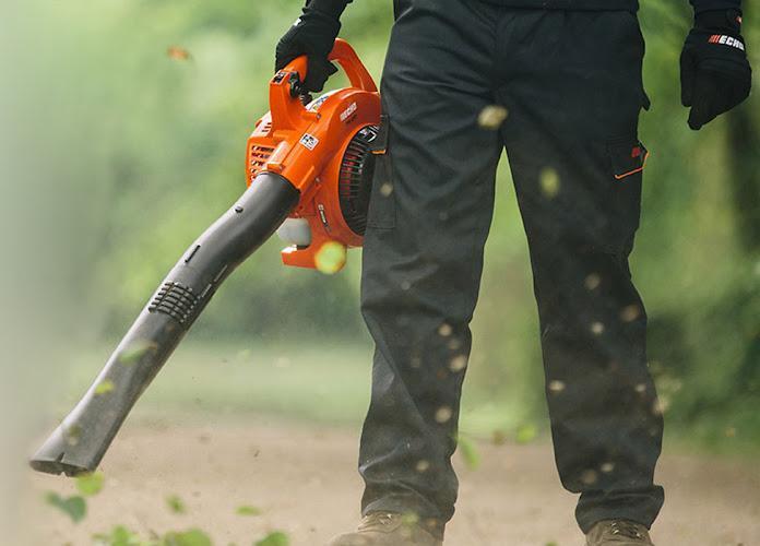 Una persona che indossa pantaloni neri e guanti aziona un soffiatore arancione, soffiando via foglie e detriti dal terreno all'esterno.