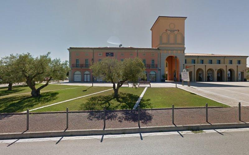 Un edificio beige e rosa con una torre dell'orologio, circondato da alberi e spazi verdi ben curati, sorge vicino a una strada asfaltata delimitata da una recinzione metallica.