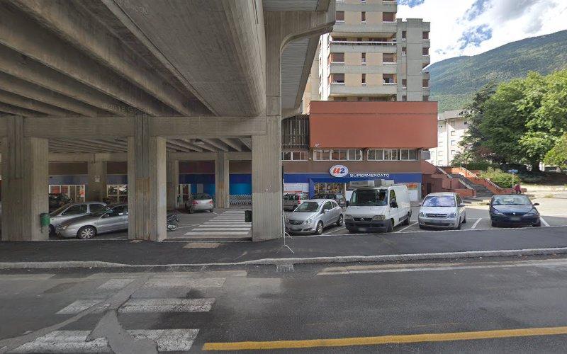 L'ingresso di un supermercato con auto parcheggiate si trova sotto una strada sopraelevata. Sullo sfondo sono visibili edifici residenziali e una montagna.