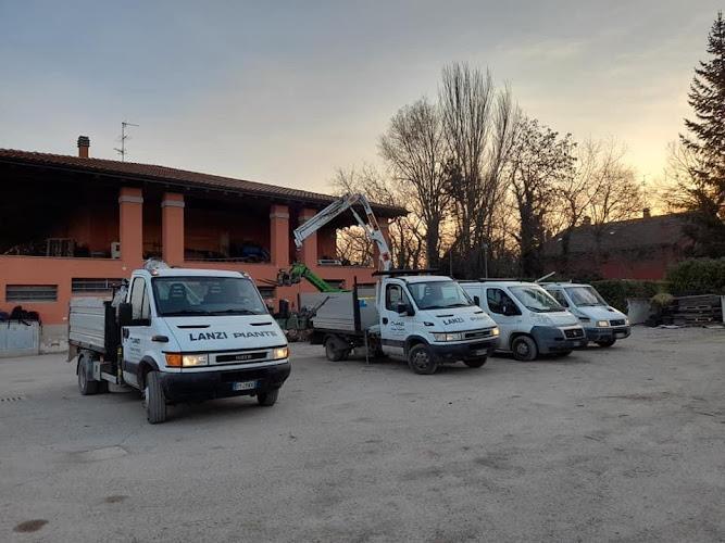 Quattro camion bianchi con il marchio "Lanzi Piante" sono parcheggiati di fronte a un edificio al tramonto. Due camion hanno le gru estese. Alberi spogli e un cielo parzialmente nuvoloso sono sullo sfondo.