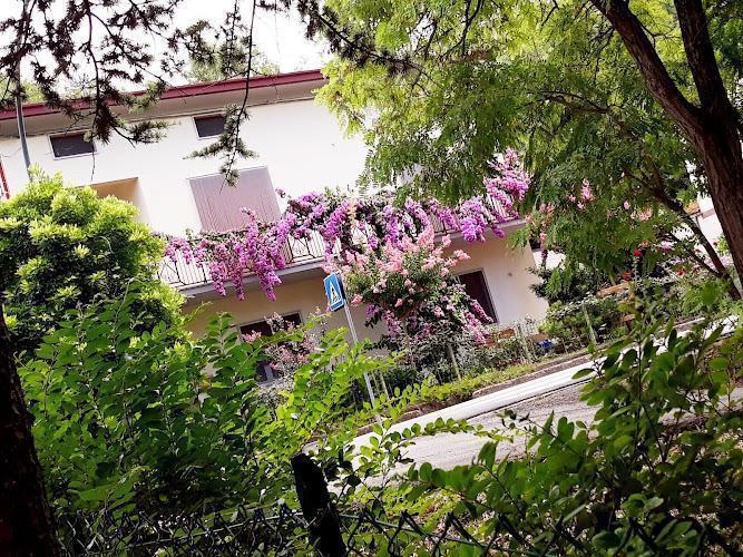 Una casa bianca a due piani con fiori viola che cadono a cascata sul balcone, circondata da rigogliosi alberi e cespugli verdi, vista da una prospettiva frontale.