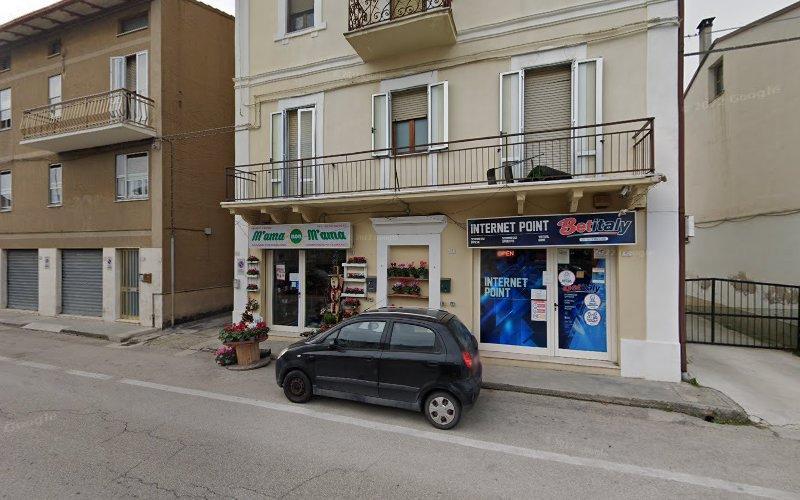 Vista stradale di un edificio di tre piani con un negozio all'angolo chiamato "Fioreria Manna" e un'adiacente vetrina "Internet Point". Una piccola auto nera è parcheggiata di fronte all'edificio.