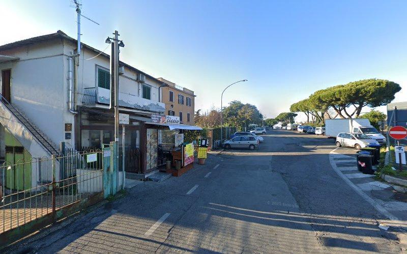 Una strada con piccoli negozi, auto parcheggiate e un pedone sul marciapiede. La strada conduce a una zona alberata. Il cielo è limpido.