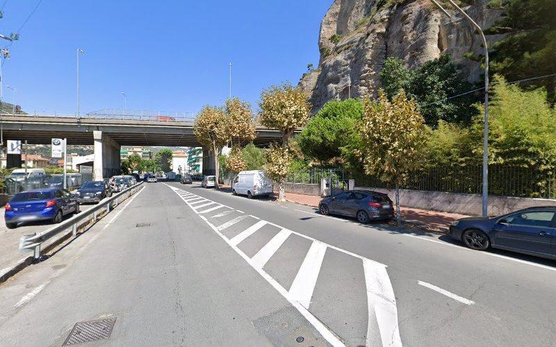 Una scena di strada con auto parcheggiate su entrambi i lati, un ponte in alto e una collina rocciosa sulla destra con alberi che costeggiano la strada. Sono visibili i cieli azzurri.