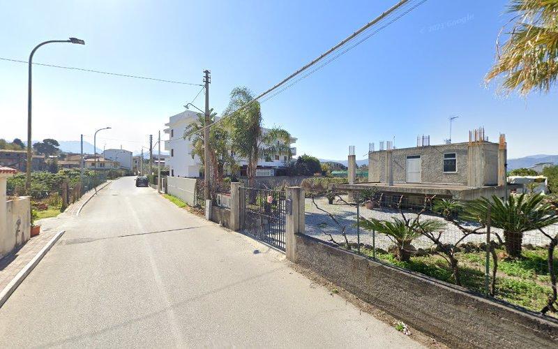 Una strada stretta fiancheggiata da case e palme in una giornata di sole. Una casa è parzialmente in costruzione, con materiali edili visibili sul posto. La strada è vuota di traffico.