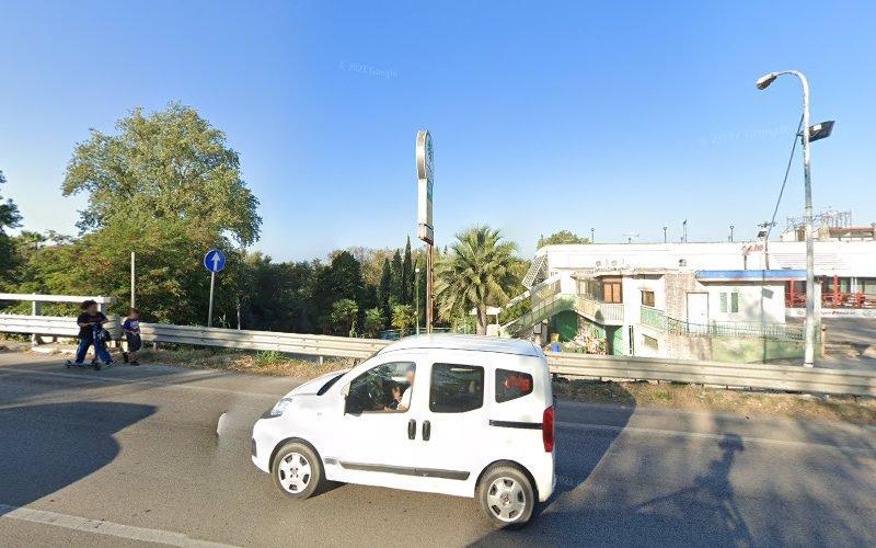 Un furgone bianco sta guidando su una strada asfaltata accanto a un edificio e ad alcuni alberi, sotto un cielo azzurro e limpido. Quattro persone stanno camminando sul marciapiede sul lato sinistro della strada.