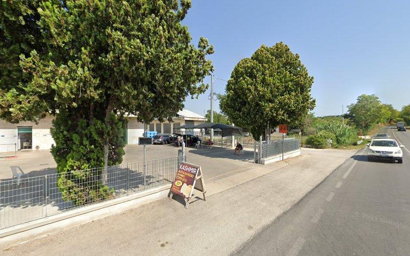 Vista stradale di un edificio con grandi alberi in primo piano, un'area recintata e auto parcheggiate. Un cartello vicino alla strada pubblicizza un prodotto o un servizio. Un'auto bianca sta passando.