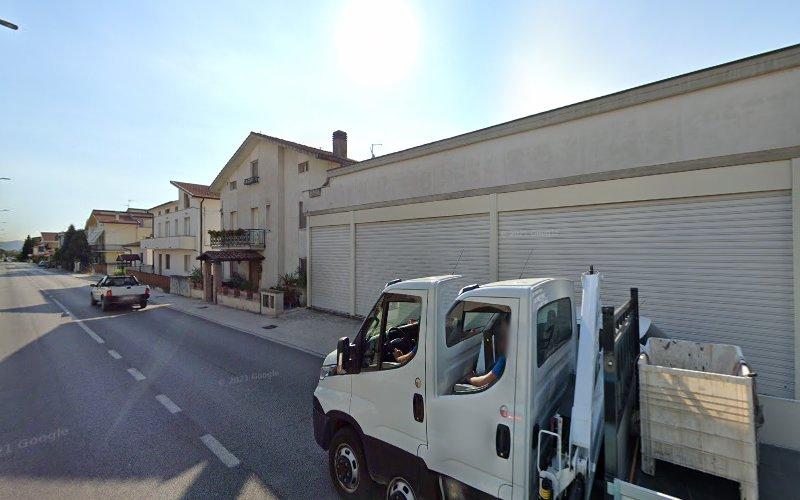 Una strada con diversi edifici, tra cui una casa con balcone e un grande magazzino con le persiane chiuse. Un camion bianco è parcheggiato sul lato destro della strada.